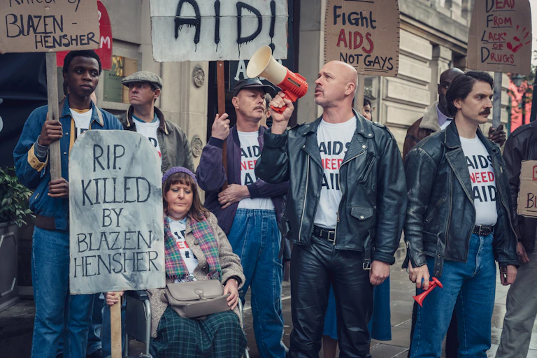 Group of people protesting holding signs that say "RIP. KILLED BY BLAZEN HENSHER", "AID", "Fight AIDS NOT GAYS" and "DEAD FROM LACK OF DRUGS"