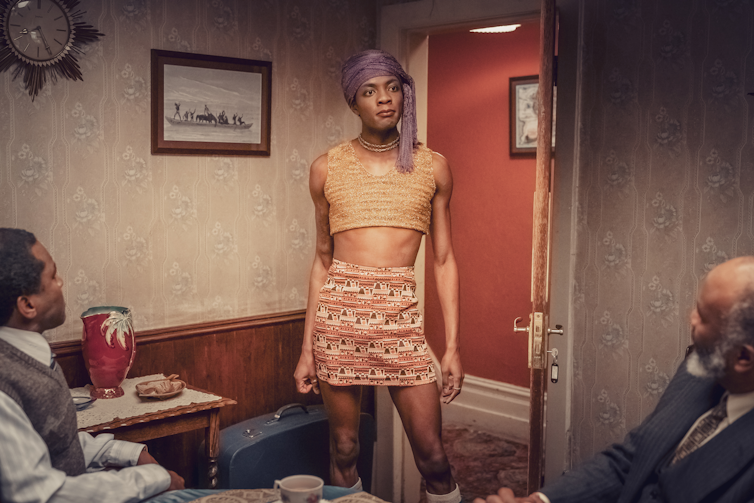 Young man wearing a beige crop top and a patterend skirt standing in a room before two suited older men