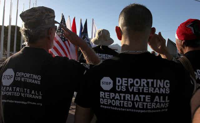 Men wearing shirts advocating for deported veterans are pictured from behind, saluting an American flag