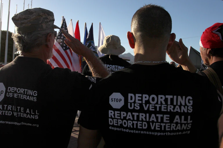 Men wearing shirts advocating for deported veterans are pictured from behind, saluting an American flag