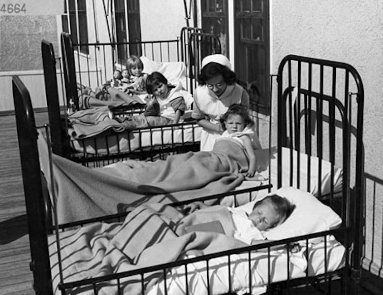 Black and white photograph of children in a row of hospital beds with an attending nurse.