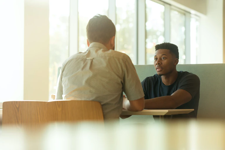 Two men sit at a table talking in an office setting.