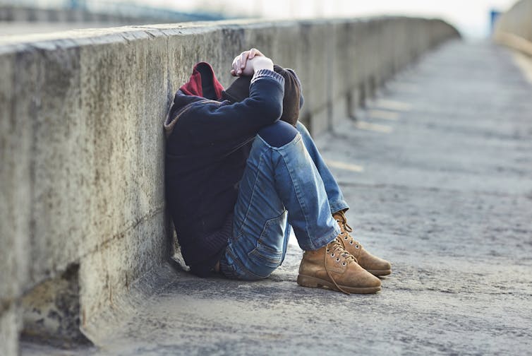 A homeless youth holds their head while sitting on the ground.