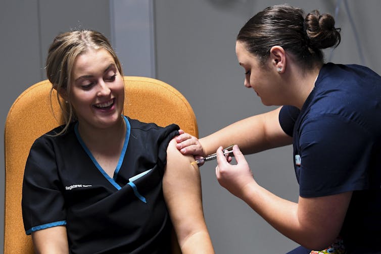 A smiling nurse receives the COVID vaccination from a fellow health-care worker.