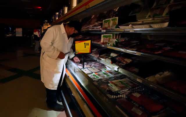 A man uses light from a cell phone to arrange products in the meat section of a grocery store.