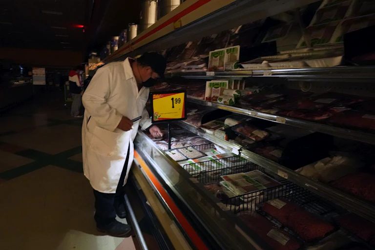 A man uses light from a cell phone to arrange products in the meat section of a grocery store.