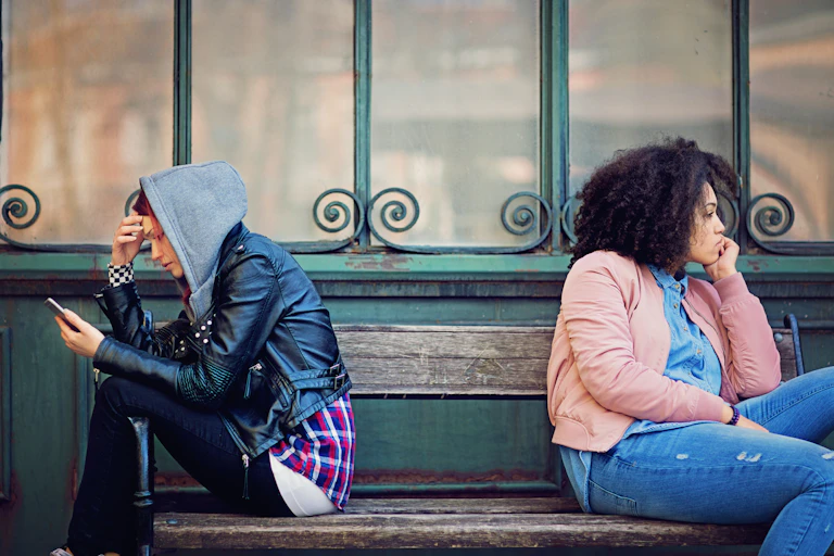 Two women sit on a bench facing away from each other, clearly upset.