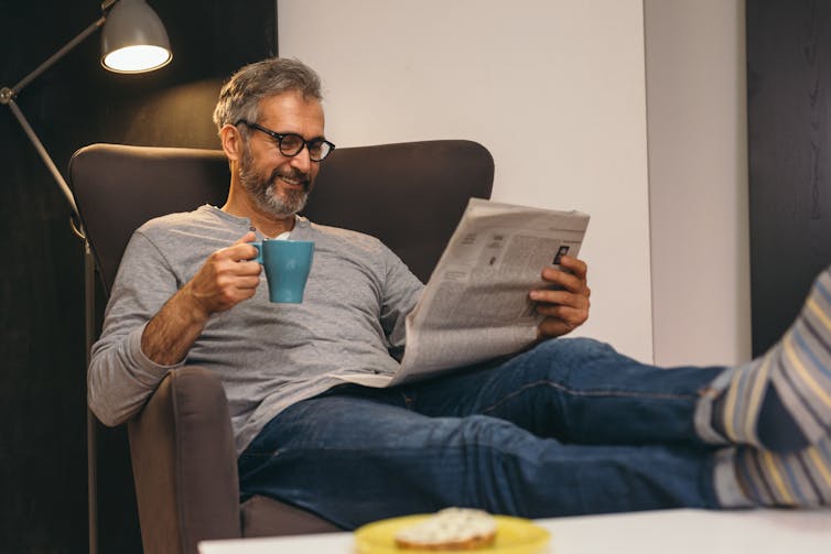 A man sits on the couch reading a newspaper, with a mug in hand.