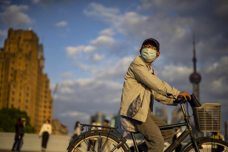 A man wearing a COVID mask rides a bicycle in with a cityscape behind him.