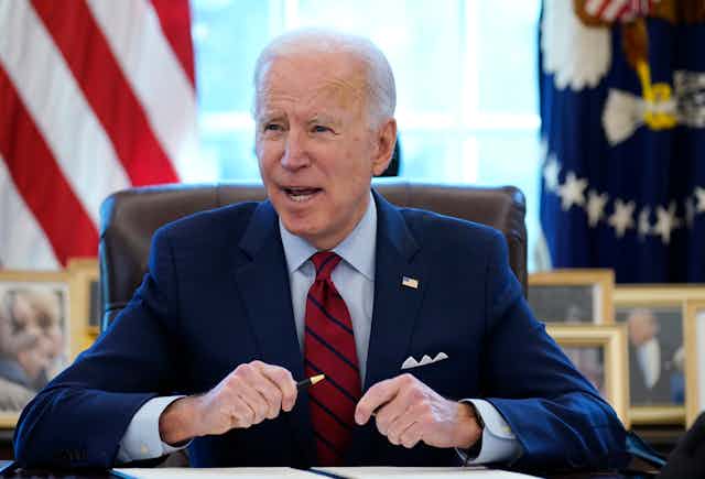 Joe Biden sits at his desk in the Oval Office holding a pen in front of a folder with papers