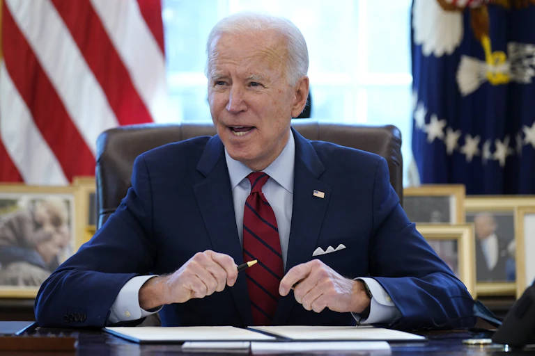 Joe Biden sits at his desk in the Oval Office holding a pen in front of a folder with papers