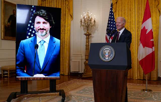 Biden stands behind a podium as Trudeau smiles on a screen.