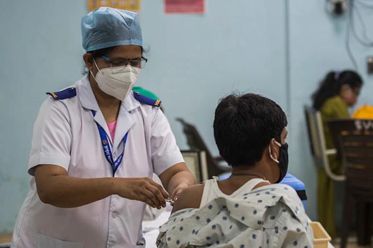 Woman in nurse's uniform, cap and face mask injects bare arm of a person also wearing a face mask