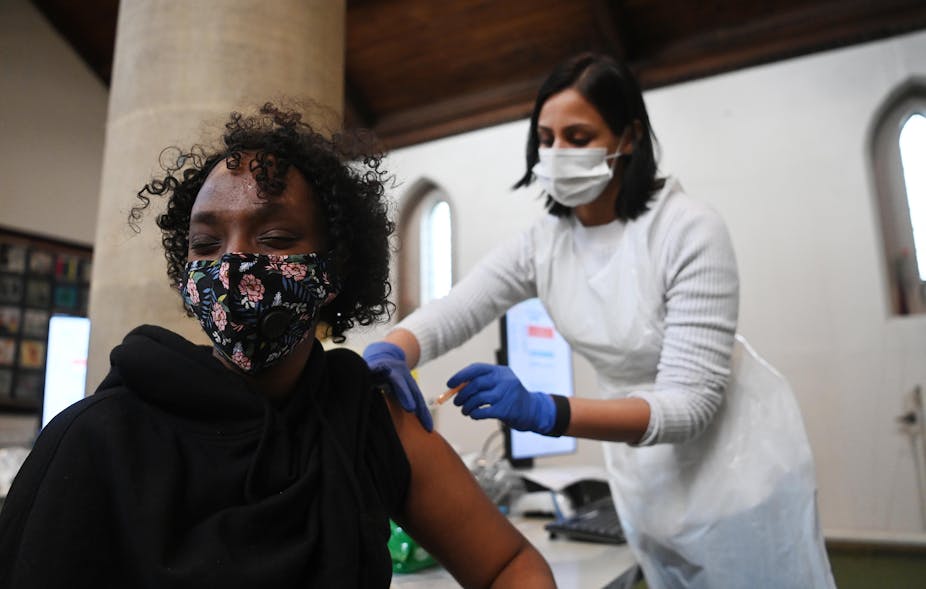 Woman in black top and face mask closes eyes while receiving the AstraZeneca COVID-19 vaccine