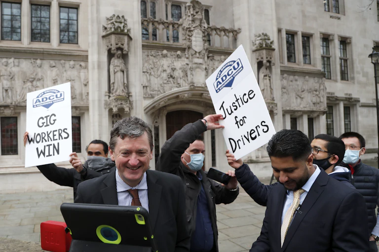Men carrying signs reading Gig Workers Win and Justice for Drivers smile outside a courthouse.