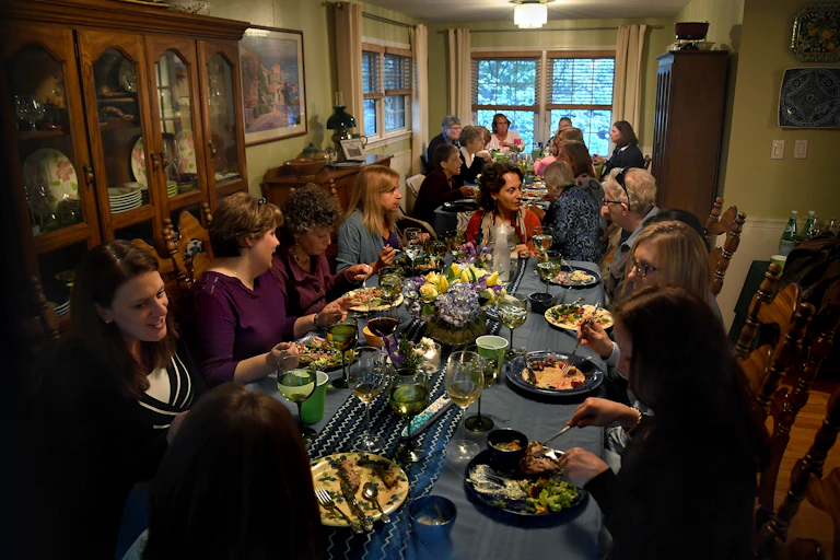 Large group of women converse and eat a shared meal at two dining room tables.