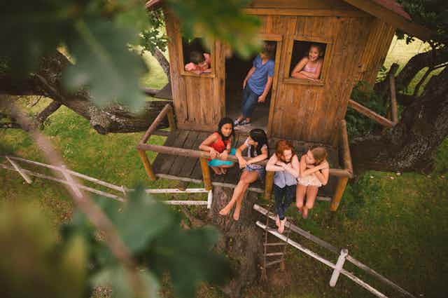 boys and girls playing and talking together in a rustic wooden treehouse, seen through the lush green leaves of a tree