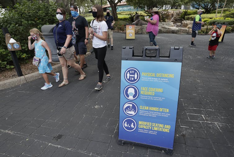 A family wearing masks walking past a sign about social distancing, mask-wearing and hand-washing.