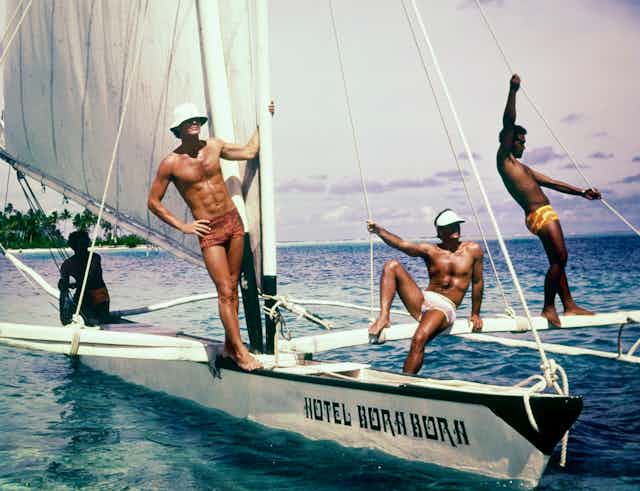 Three models pose on a sailboat wearing swim trunks.