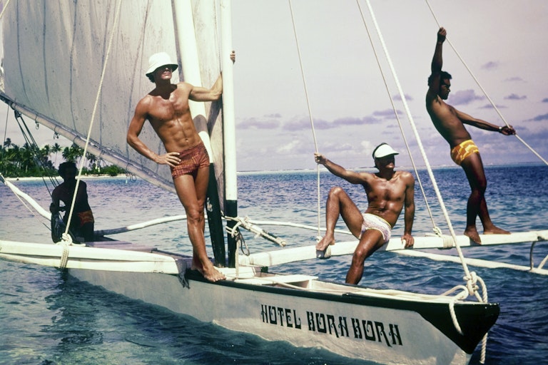 Three models pose on a sailboat wearing swim trunks.