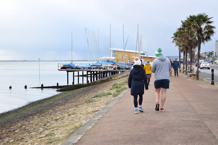 Man wearing shorts and woman wearing winter coat walking near the seaside in cold weather.