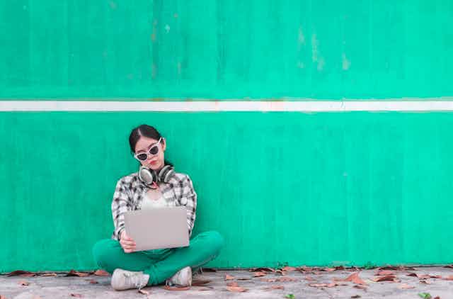 A teenager sitting against a green wall with her laptop in front of her.