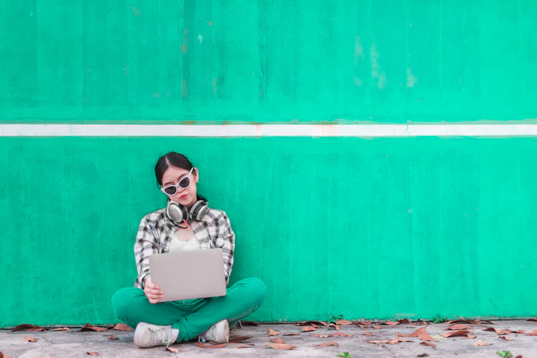 A teenager sitting against a green wall with her laptop in front of her.