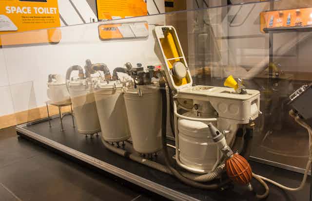 A display of a space toilet showing funnel, suction devices and four tanks to store waste.