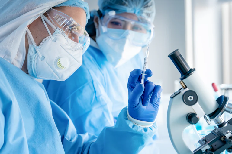 Two scientists in PPE looking at syringe, microscope