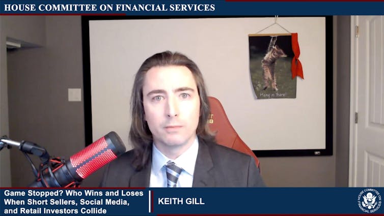 Screenshot of video call featuring man wearing a suit, with poster of a cat and caption 'Hang in there' in the background.