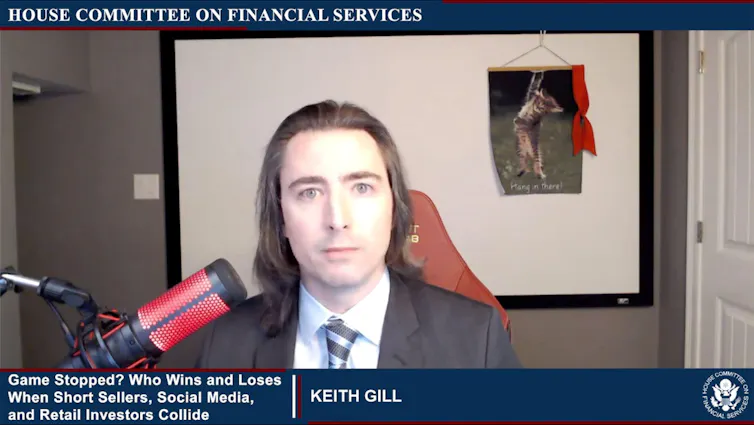 Screenshot of video call featuring man wearing a suit, with poster of a cat and caption 'Hang in there' in the background.