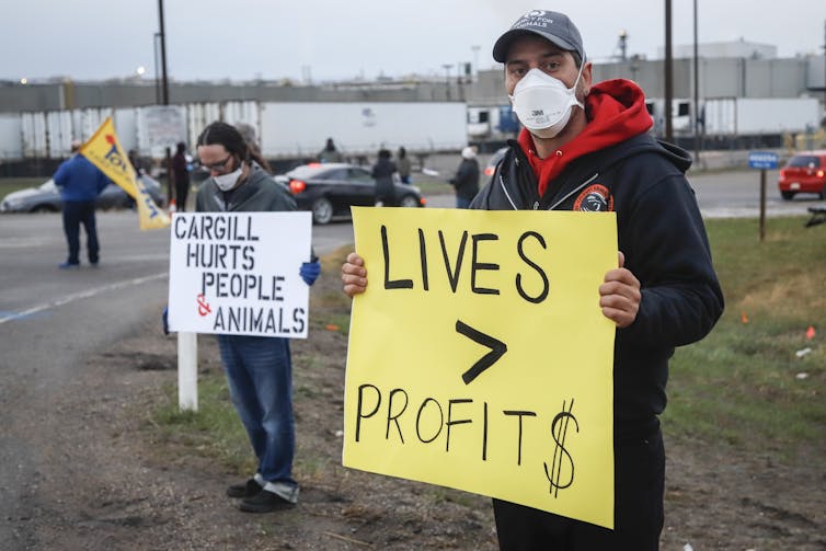 Protesters stand on the side of the road carrying a Lives Before Profits sign