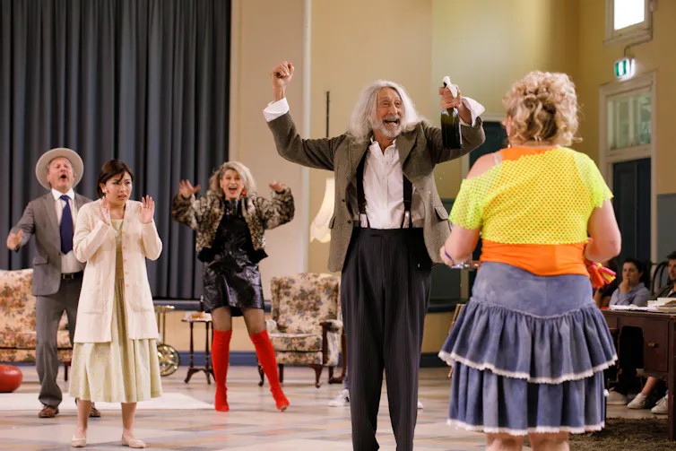 Production image: the cast parties in a hall