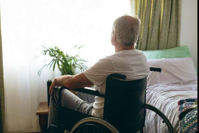 A senior man sits in a wheelchair next to a bed, looking out the window.