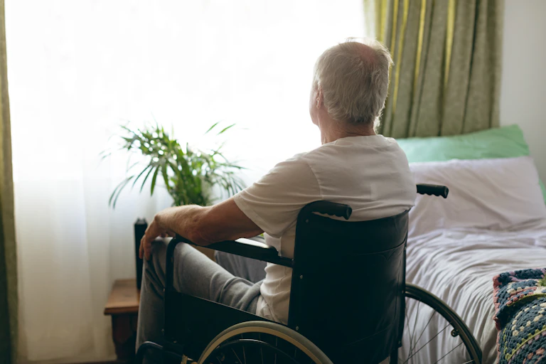 A senior man sits in a wheelchair next to a bed, looking out the window.