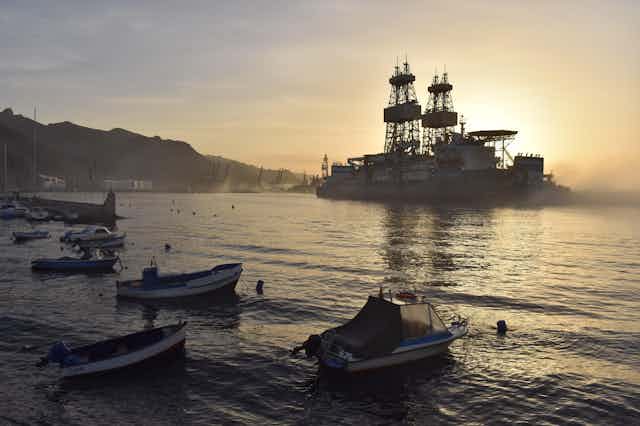 A drilling platform looms large in a calm bay with small boats in foreground.