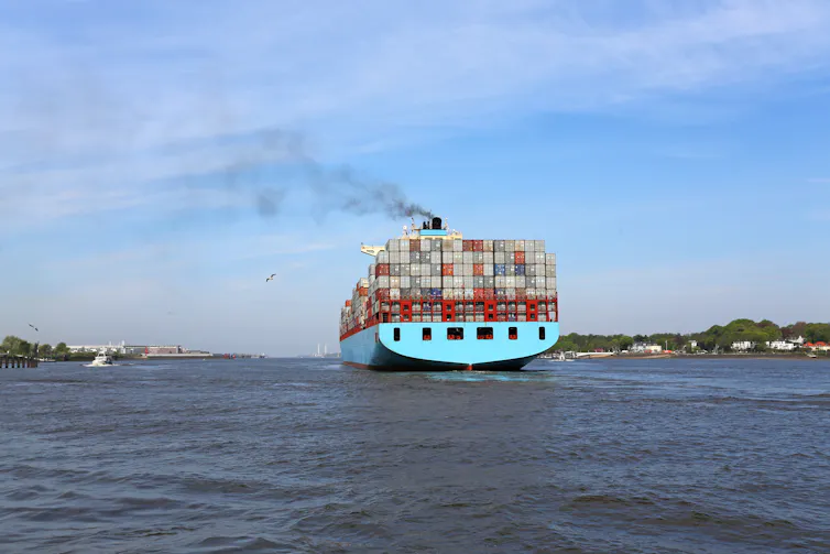 A large container ship leaving port belches black smoke.