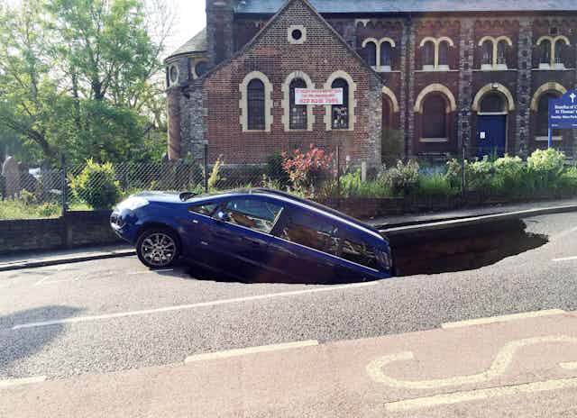 A blue car falling into a sinkhole in the road. Church in background.
