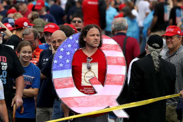 A Trump supporter at a rally wears a giant cutout 'Q.'