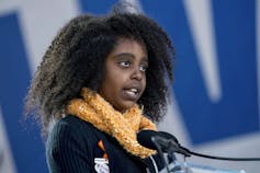 A girl wearing an orange scarf talks into a microphone.