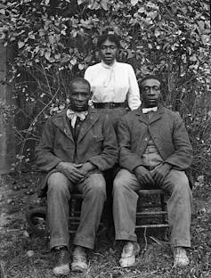 Two Black men seated in chairs while their sister stands behind them.