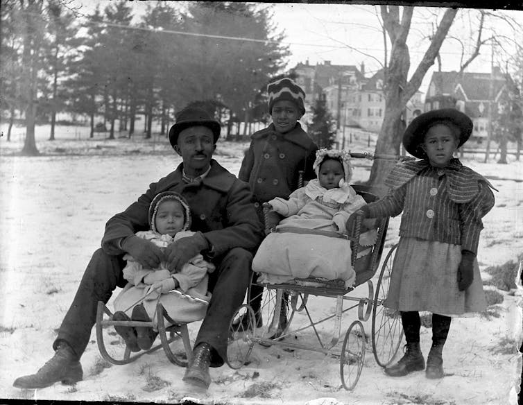 A man sits on a sled with one of his children, while another is in a baby carriage and two stand.
