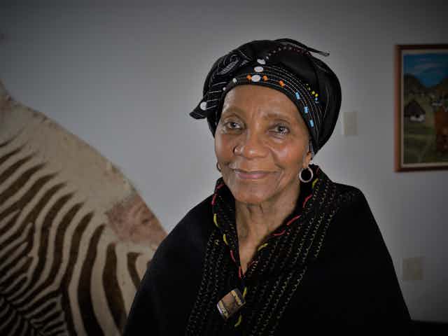 An elder woman smiles lightly as she looks into the camera, eyes warm and dressed in black, beaded traditional Xhosa attire, a zebra skin on the wall behind her.
