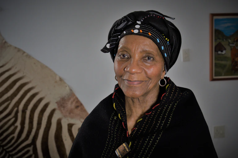An elder woman smiles lightly as she looks into the camera, eyes warm and dressed in black, beaded traditional Xhosa attire, a zebra skin on the wall behind her.