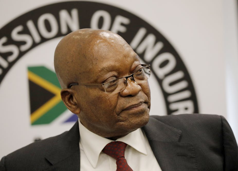 A man with a shiny baldhead wearing specs, a white shirt, red tie and a black dark jacket sits on a chair with his back to the emblem of South Africa’s commission investigating grand corruption.