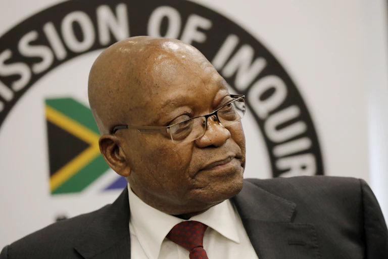 A man with a shiny baldhead wearing specs, a white shirt, red tie and a black dark jacket sits on a chair with his back to the emblem of South Africa’s commission investigating grand corruption.