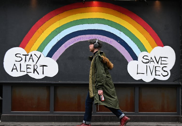 A man walks past a mural of a rainbow and two clouds saying 'Stay Alert' and 'Save Lives'