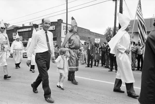 Archival image of Ku Klux Klan members walking in a parade while protesters gather on the side