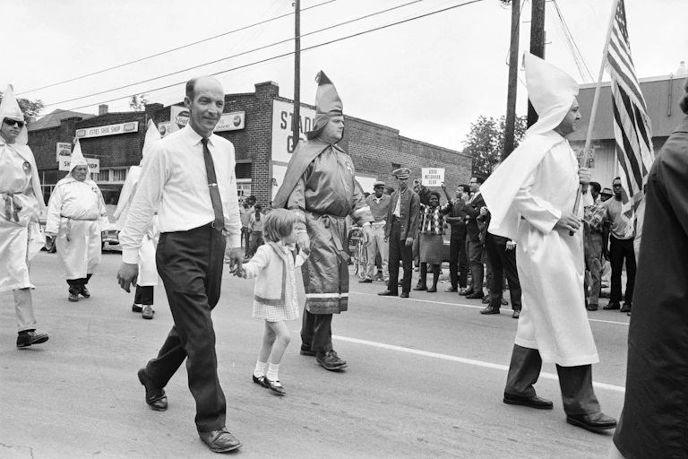 Archival image of Ku Klux Klan members walking in a parade while protesters gather on the side