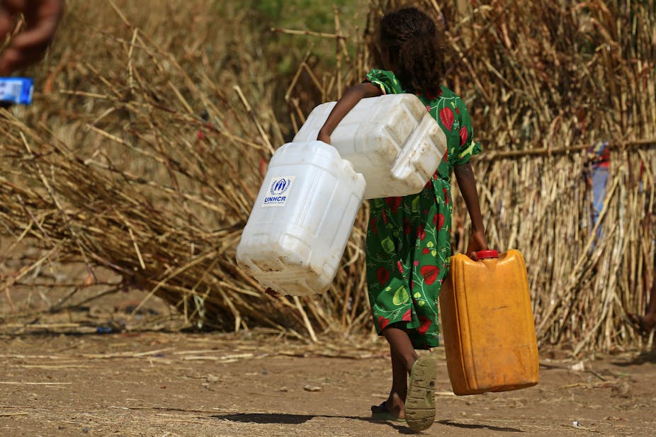 A young girl in a green dress carrying white and yellow water containers in a refugee camp